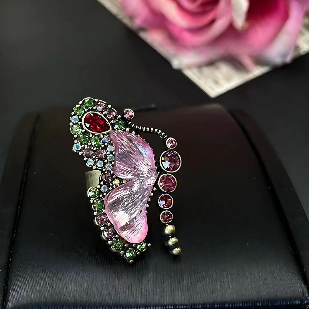 Butterfly-shaped brooch with gemstones on a black surface with a pink rose in the background