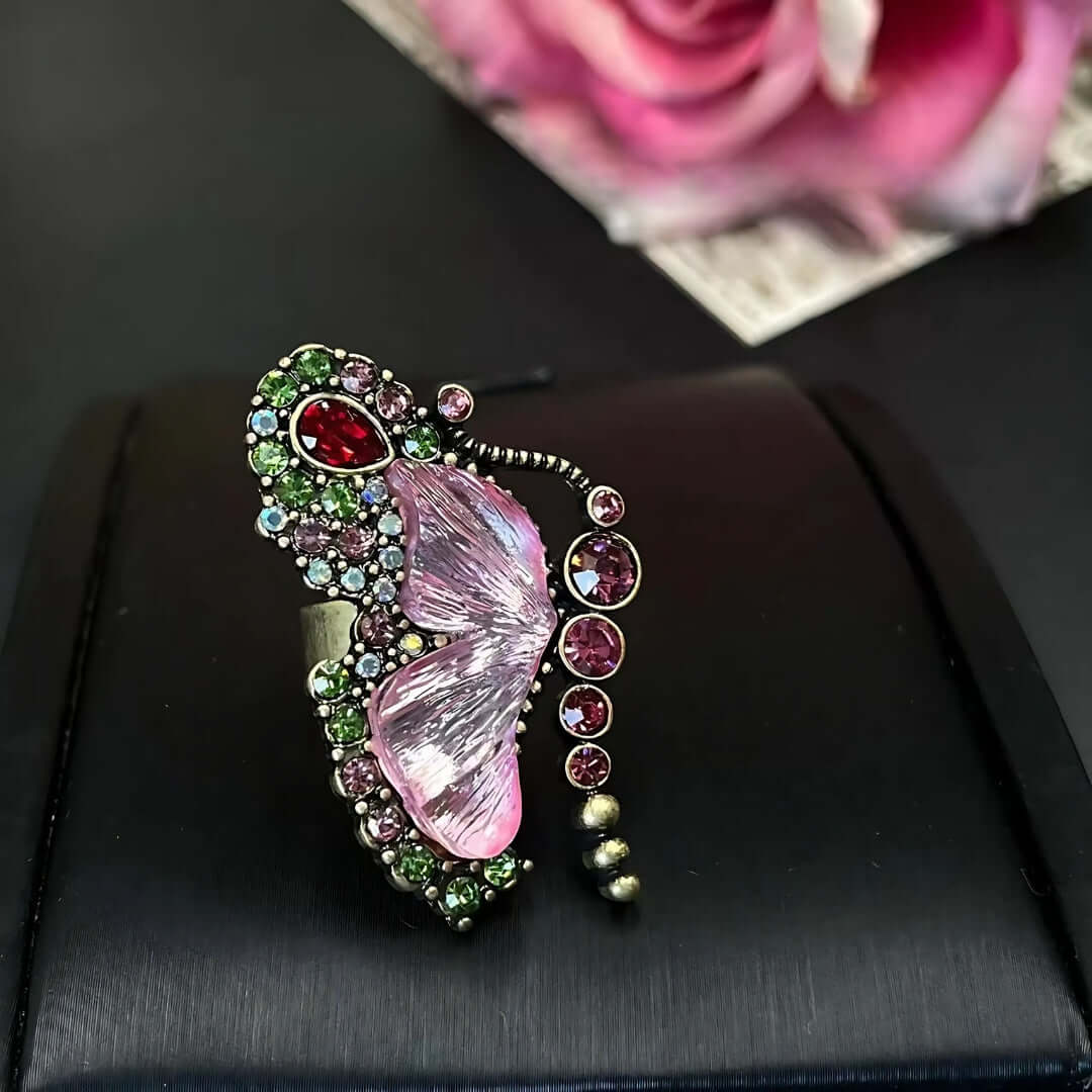 Butterfly-shaped brooch with gemstones on a black surface with a pink rose in the background