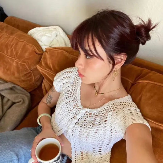 Woman sitting on a brown couch holding a cup, wearing a white crochet top, wearing jazzy tune layering necklace and hoop earrings