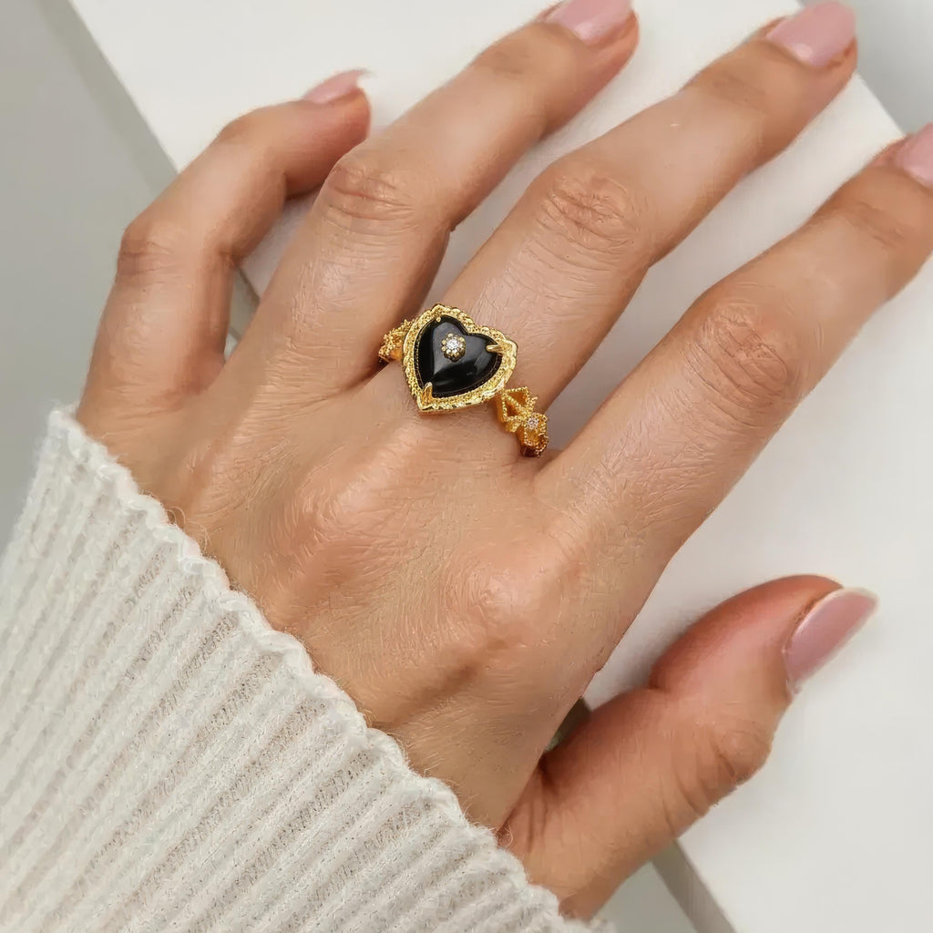 Hand wearing a gold ring with a black heart-shaped Paua Shell Ring on a neutral background