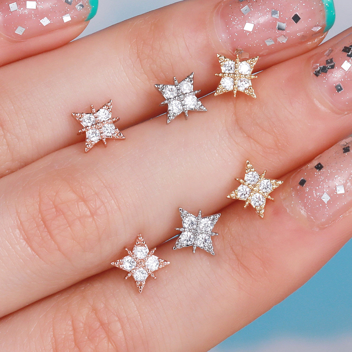 Assorted tiny star-shaped cartilage earrings in rose gold, gold, and white gold, held by a hand with glittery nail polish.