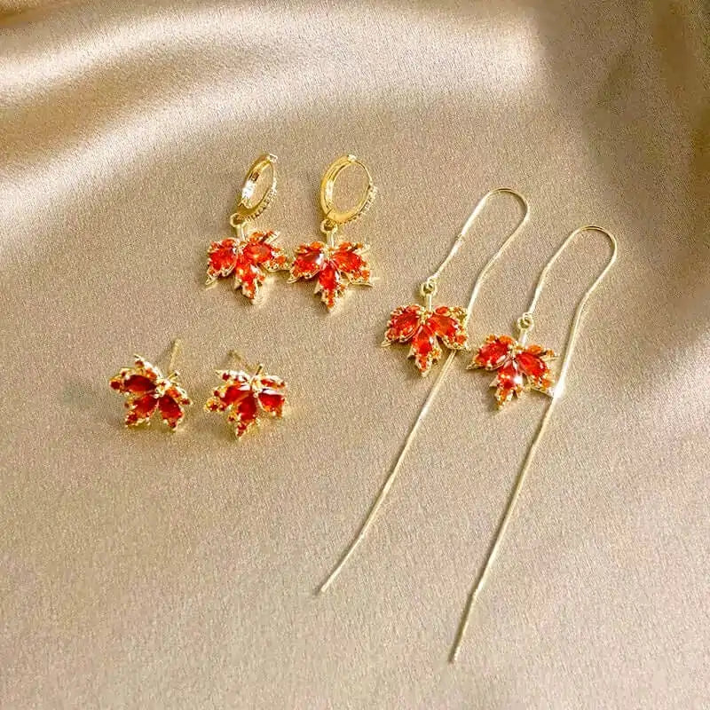 Set of gold earrings with orange maple leaf threader earrings, stud earrings and hoop earrings on a beige background