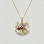 Gold necklace with a cat-shaped pendant featuring floral and fruit designs on a light gray background.