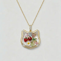 Gold necklace with a cat-shaped pendant featuring floral and fruit designs on a light gray background.