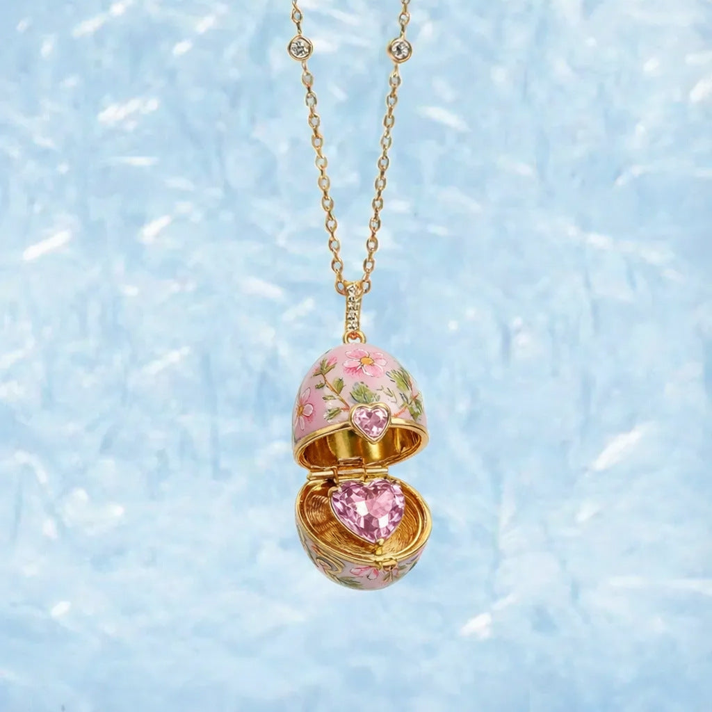 Decorative egg-shaped pendant with pink heart and floral design on a gold chain against a light blue background.